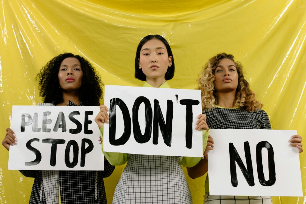 Three women stand united holding protest signs against a vivid yellow backdrop.