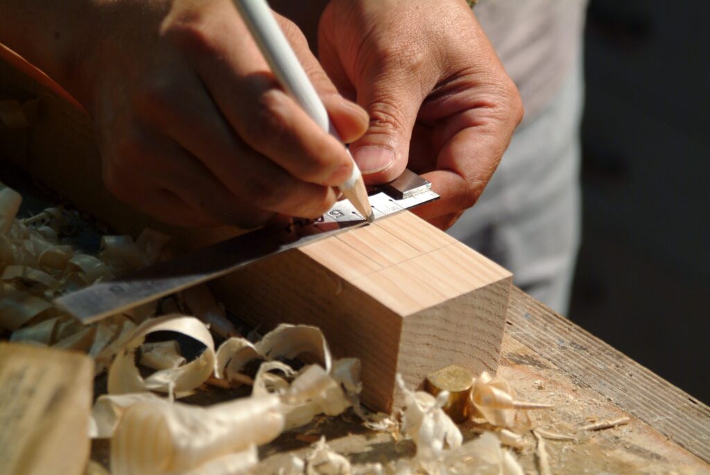 Nói Thì Dễ, Làm Đi Close-up of hands measuring a wooden plank using a ruler and pencil on a workbench.