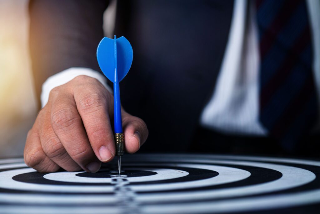 Mục Tiêu Close-up of a man's hand holding a blue dart on a bullseye, symbolizing precision and focus.